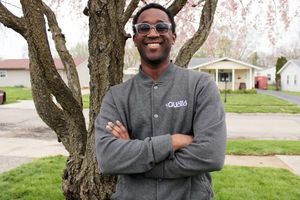 Christian smiling outdoors in a grey jacket with a small You Are A CEO logo on the chest
